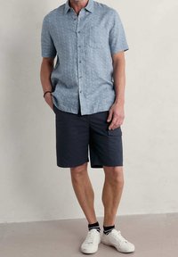 Korte mouwen, lichtblauw shirt met een geometrisch patroon, gecombineerd met donkere marineblauwe shorts. Witte sneakers met zwart-witte gestreepte sokken.