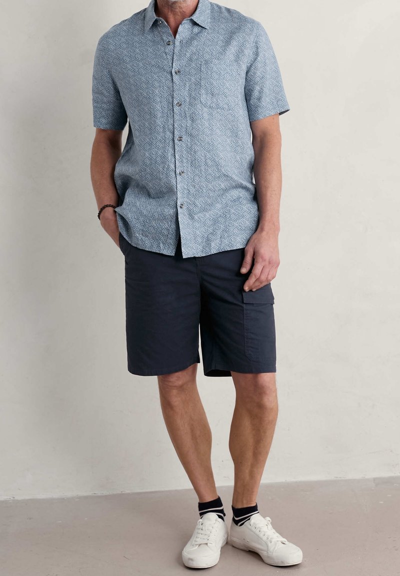 Korte mouwen, lichtblauw shirt met een geometrisch patroon, gecombineerd met donkere marineblauwe shorts. Witte sneakers met zwart-witte gestreepte sokken.
