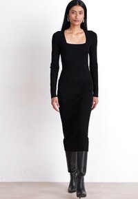 Black form-fitting long-sleeve dress with a square neckline, made of textured fabric. Model wears knee-high black boots for contrast.