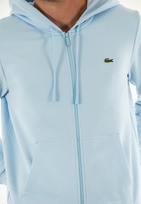 Sudadera azul con cremallera hecha de una tela suave, que cuenta con capucha, bolsillos frontales y un pequeño logo verde bordado en el área del pecho izquierdo.