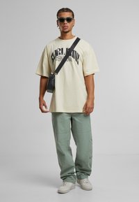 Tejfehér, nagyméretű t-shirt grafikával, világoszöld cargo nadrággal, fekete keresztpántos táskával és fehér sportcipővel. Strukturált anyag és laza fit.