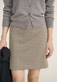 Jupe à carreaux marron et beige avec un design ajusté, associée à un cardigan en tricot gris. La jupe arrive au milieu de la cuisse. Tissu noir drapé dans la main gauche.
