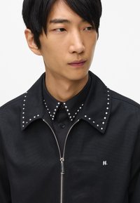 Melna jaka ar rāvējslēdzēju priekšā, ar apkakli, kas rotāta ar sudraba kniedēm un mazu logotipu uz krūtīm. Apdare uz vienkrāsaina, teksturēta auduma.