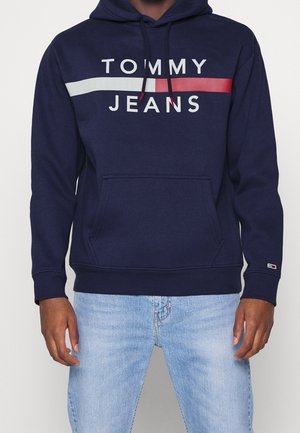 Mężczyzna w granatowej bluzie Tommy Jeans z białym i czerwonym paskiem, w połączeniu z jasnoniebieskimi dżinsami, stojący na jednolitym tle.