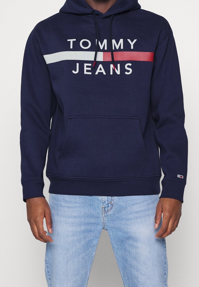 Homme portant un sweat à capuche Tommy Jeans bleu marine avec une bande blanche et rouge, associé à un jean bleu clair, debout devant un fond uni.