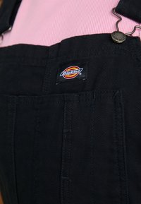 Črne platnene naramnice z nastavljivimi trakovi, sprednjim žepom in vidno etiketo Dickies. Tkanina ima teksturiran zaključek.