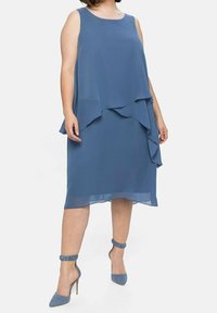 Robe en chiffon bleue avec un design en couches, sans manches et une longueur au genou. Associée à des escarpins bleus assortis.