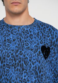 Zila leoparda raksta džemperis ar melnu sirdīšu aplikāciju uz krūšu daļas. Audumam ir teksturēta virsma un apaļš izgriezums.