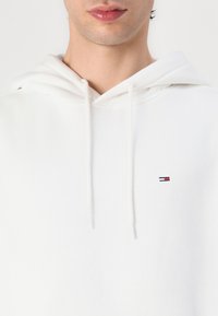 Haut à capuche blanc avec cordon de serrage, arborant un petit logo brodé en rouge, blanc et bleu. Texture lisse, en molleton de moyenne épaisseur.