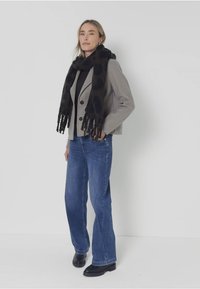 Blazer grigio sopra un dolcevita nero, abbinato a jeans a zampa d'elefante blu e a una sciarpa nera con macchie. La sciarpa ha bordi a frange.