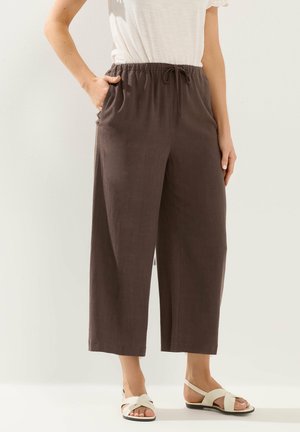 Kobieta w luźnych ciemnobrązowych spodniach o długości 7/8 z elastycznym paskiem i białych sandałach, stojąca na tle jasnej ściany.