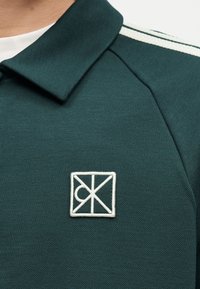 Σκούρο πράσινο πόλο με γιακά, με λευκή ετικέτα λογότυπου και δύο λευκές ρίγες στον ώμο, φορεμένο πάνω από ένα λευκό μπλουζάκι.