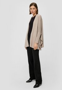 Beige open cardigan met een losse pasvorm, zachte textuur en verlaagde schouders, uitgevoerd met zwarte, op maat gemaakte broek en puntige zwarte schoenen.