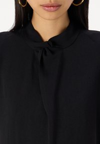 Schwarze Bluse mit rundem Ausschnitt, die an der Vorderseite mit einer gebundenen Schleife versehen ist. Hergestellt aus weichem Stoff mit glatter Textur und minimalistischem Design.