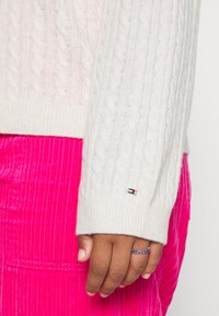 Main avec bague en pierre violette portant un pull blanc en maille torsadée et un pantalon en velours côtelé rose vif, gros plan de côté.