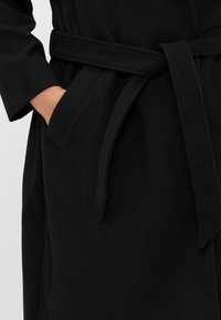 Musta viitta, joka on valmistettu pehmeästä kankaasta ja jossa on vyötärölle sidottava vyö, syvät etutaskut ja sileä tekstuuri. Muotoillut hihat, joissa on rento istuvuus.