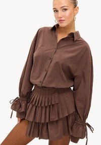 Chemise marron à boutons, manches longues avec poignets froncés, associée à une mini-jupe à volants à taille ajustée et à plis.