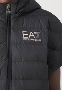 Černá vycpaná vesta s zipovým uzávěrem, kapucí a stříbrným logem "EA7 EMPORIO ARMANI" na přední straně. Hladký povrch.