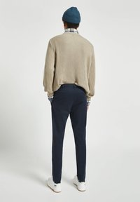 Un pull beige à côtes, un pantalon bleu marine et des baskets blanches. Le pull a un col rond et des manches bouffantes, avec une chemise à carreaux bleue et blanche qui dépasse.