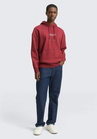 Maronenfarbener Kapuzenpulli mit einer Vorderentasche und Kordeln, kombiniert mit dunkelblauen Jeans und weißen Turnschuhen.