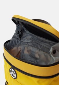 Zaino giallo con un'esterno liscia e impermeabile. L'interno presenta una fodera grigia con tasche, chiusura con zip e dettagli con bottone a pressione.