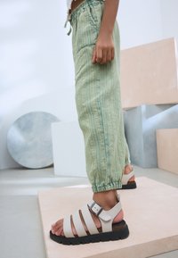Light green, textured, elasticized pants with vertical stripes paired with white, open-toed sandals featuring three straps and a buckle.