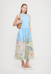 Abito senza maniche di colore azzurro chiaro con un busto aderente e una gonna ampia e decorata che ritrae un paesaggio colorato. La modella tiene una borsa intrecciata.