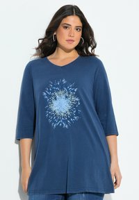 Frau mit dunklen Haaren, die Ohrringe in Form von Ringen trägt und ein blaues Tunika-Oberteil mit einem zentralen floralen Punktdesign an hat, steht vor einem weißen Hintergrund.