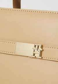 Détail d'un sac à main en cuir beige avec une sangle cousue et un fermoir rectangulaire doré arborant un logo "TH" entrelacé.