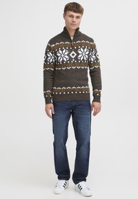 Maglione lavorato a maglia marrone con motivo a fiocchi di neve bianco e arancione, colletto con zip, abbinato a jeans blu e sneakers bianche.