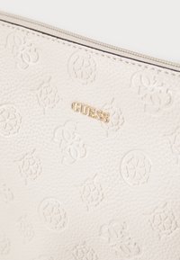 Portefeuille en cuir blanc texturé avec motifs floraux et logo en relief, présentant un petit nom de marque "GUESS" en métal doré sur le devant.
