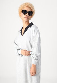 Vestido de suéter oversized cinza com um decote colorido preto e detalhes em botão. Manga longa e um ajuste descontraído. Sem padrões visíveis.