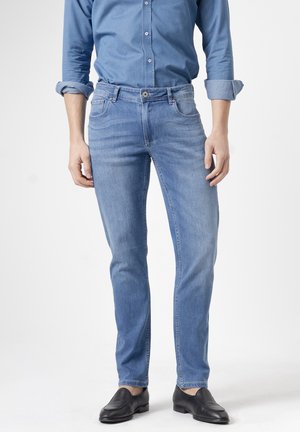 Persona con vaqueros ajustados azul claro, camisa azul abotonada con las mangas remangadas y zapatos negros sin cordones, de pie frente a un fondo blanco.