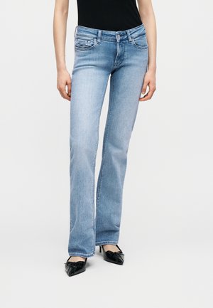 Moteris dėvi šviesiai mėlynas aptemptas džinsines kelnes su platėjančiais apačioje plėtais, juodus smailianosius aukštakulnius su lankeliais ir juodą be rankovių viršutinę dalį, stovinčią priešais paprastą foną.