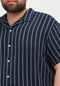Camisa de manga corta abotonada en tejido azul marino oscuro con rayas verticales en blanco y azul claro, y un diseño de cuello relajado.