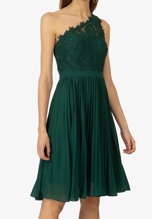 Kobieta w ciemnozielonej sukience na jedno ramię o długości do kolan, z koronkowym gorsetem i plisowaną spódnicą z szyfonu, na białym tle.