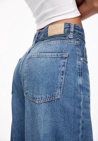 Blå denimjeans med hög midja, försedd med en beige läderlapp bak, standard bakfickor och en avslappnad passform.