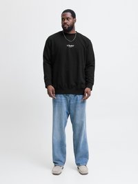 Sudadera negra con logo blanco, combinada con jeans de pierna ancha en azul claro y zapatos tipo slip-on beige. Fondo liso, estilo casual.