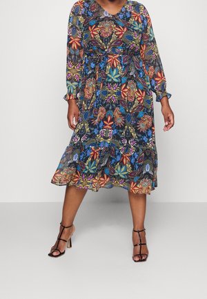 Frau, die ein buntes Midikleid mit Blumenmuster, langen Ärmeln und schwarze, Riemchen-hochabsatzsandalen trägt, steht vor einem schlichten Hintergrund.