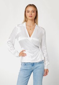 Balta satino palaidinė su ilgomis rankovėmis, apykakle ir priekine užrišimo detale, derinama su šviesiai mėlynomis džinsinėmis kelnėmis. Lygi tekstūra, elegantiškas dizainas.