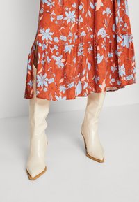 Beige kniehohe Stiefel mit glänzender Oberfläche, spitzer Zehenpartie und Blockabsatz, kombiniert mit einem rot gemusterten Kleid mit blauen Blumen.
