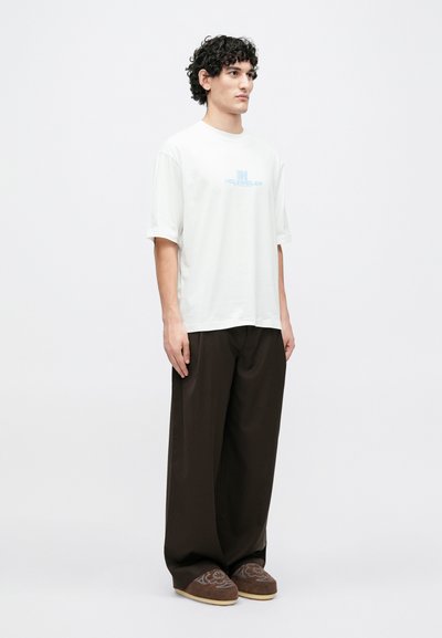 Tricou alb cu mâneci scurte și lejer, cu logo-ul "HOLZWEILER" în albastru deschis, asortat cu pantaloni largi maro și pantofi slip-on maro cu model.