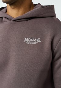 Barna kapucnis pulóvert viselő férfi, amelyen fehér szöveg: "CLSR LDN Luxury British Atelier" az egyik mellkasán, vállaktól felfelé fényképezve.