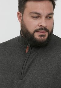 Tamno sivi dukserica sa patentnim zatvaračem i rebrastim ovratnikom. Ima mali kožni držač za patentni zatvarač. Mekani, teksturirani pleteni materijal.