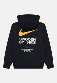 Crna dukserica s velikim narančastim Nike swoosh logotipom na leđima, bijelim tekstom i kapuljačom svezanom sa špagom. Izrađena od mekanog, teksturiranog materijala.