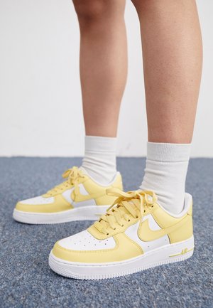 Piernas con calcetines blancos hasta el tobillo y zapatillas Nike Air Force 1 amarillas y blancas, de pie sobre una alfombra azul.