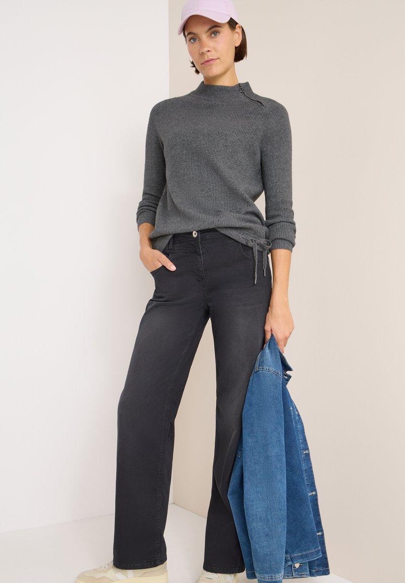 Grijze geribde trui met een zijkraag, zwarte broek en een spijkerjack in de hand. Een lichtroze pet en neutrale sneakers maken de look compleet.