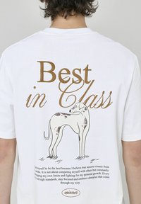 T-shirt en coton blanc avec un design imprimé d'un chien et le texte "Meilleur de sa classe" en marron. Inclut un texte motivant et le nom de la marque "winner".
