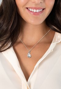 Silverhalsband med en snirklig design och en rund pärlprydnad, visad på en kedja mot en ljus blus.