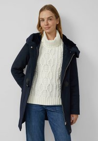 Frau trägt einen marineblauen Kapuzenmantel über einem weißen Strickpullover mit Rollkragen und blauen Jeans, steht vor einem schlichten hellen Hintergrund.